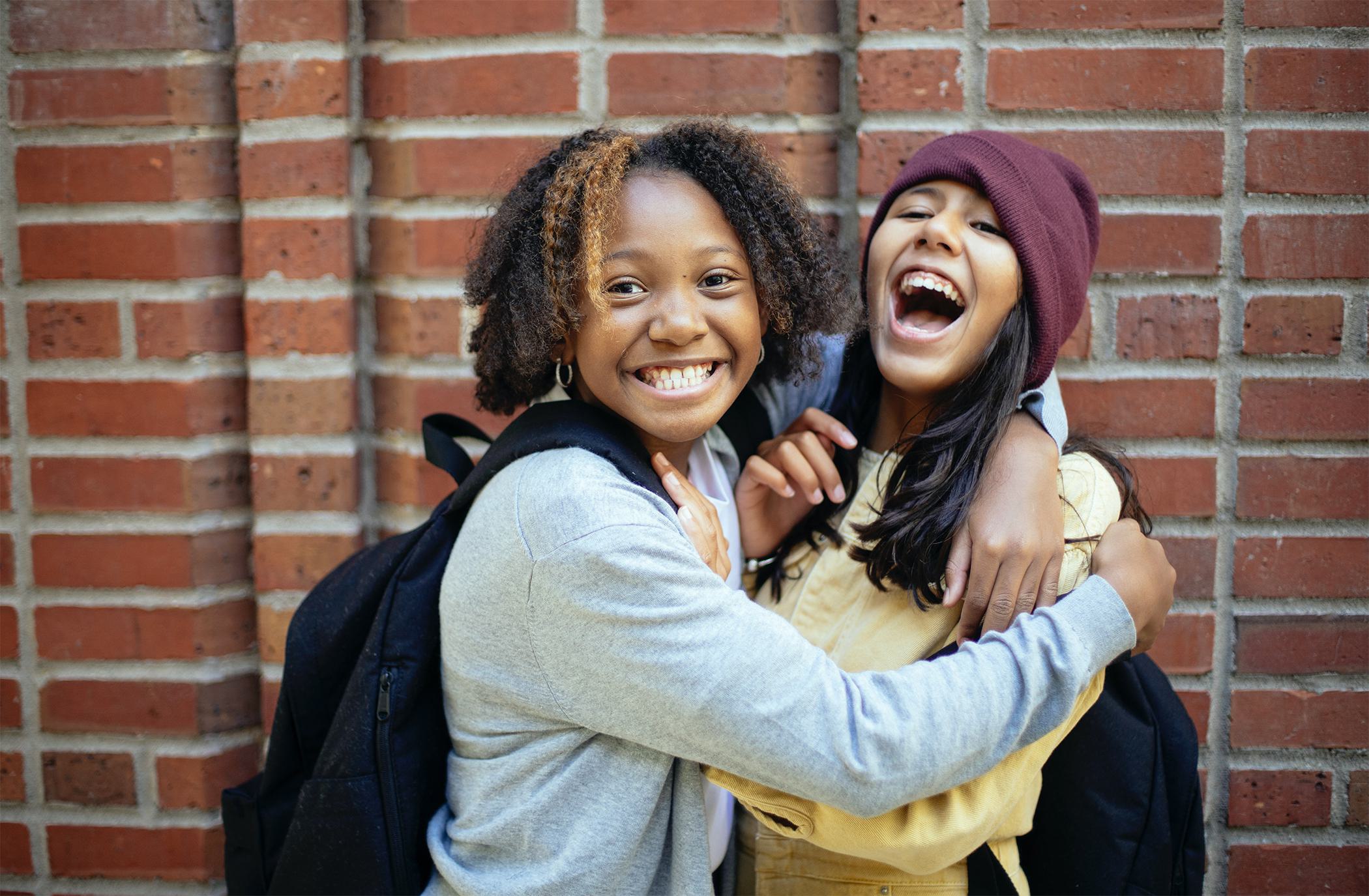 Two girls with backpacks that are hugging and laughing in front of a red brick wall.